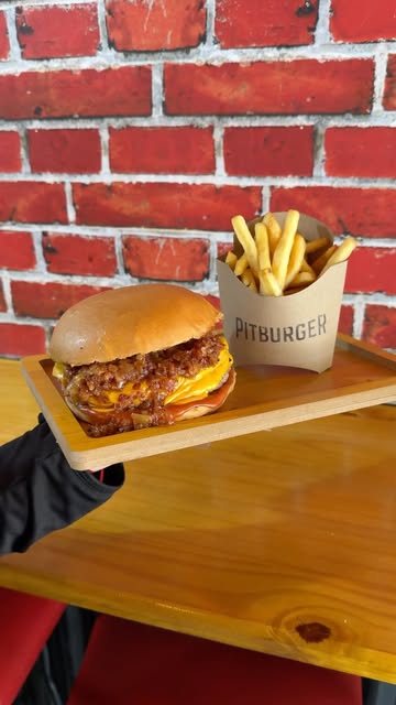 Hamburguesa doble con cheddar y bacon y papas fritas Pitburger sobre tabla de madera, con pared de ladrillo detrás